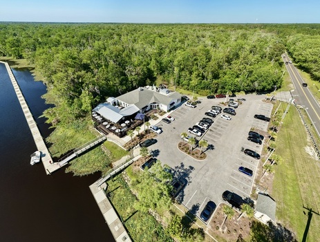 Aerial view of Sixmile Creek featuring a waterfront restaurant with a parking lot and a long wooden pier surrounded by dense green forest in St. Johns County Florida United States.