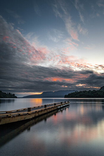 View of Lake Tarawera with a wooden jetty extending into the calm water under a dramatic sunset sky with clouds and distant mountains in Rotorua Bay of Plenty Region New Zealand.