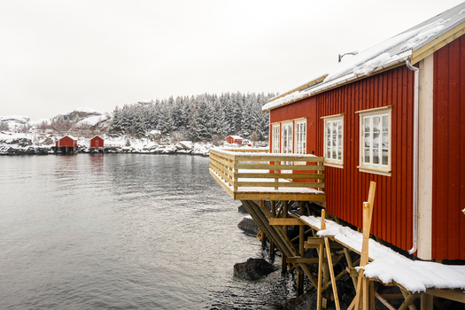 View of a traditional red wooden cabin on stilts over the calm water with snow covered trees and hills in the background in Leknes Lofoten Norway.