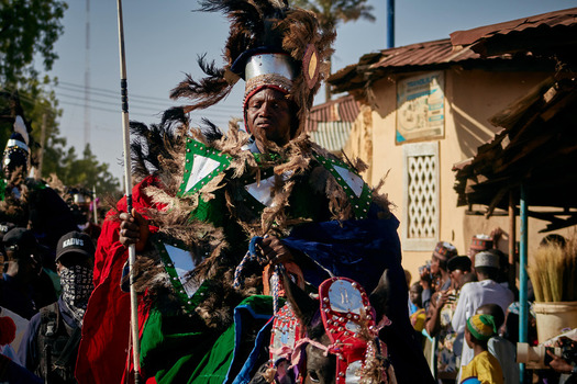 Zaria Nigeria 22 March 2026 View of a regal figure in a vibrant feathered costume and gleaming headpiece holding a spear amidst the bustling street. Zaria Nigeria 22 March 2026 View of a regal figure in a vibrant feathered costume and gleaming headpiece holding a spear amidst the bustling street.