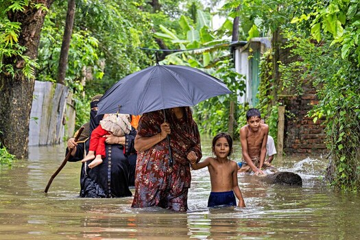 Banshkhali Upazila Chittagong Division Bangladesh 10 July 2026 View of a family wading through floodwaters in a rural village with a woman holding an umbrella and children walking. Banshkhali Upazila Chittagong Division Bangladesh 10 July 2026 View of a family wading through floodwaters in a rural village with a woman holding an umbrella and children walking.