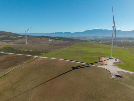 Aerial view of modern wind turbines standing in green agricultural fields with distant mountains under a clear blue sky in Swellendam Western Cape South Africa. Aerial view of modern wind turbines standing in green agricultural fields with distant mountains under a clear blue sky in Swellendam Western Cape South Africa.