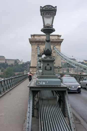 View of the ornate lamppost and steel structure of the historic Szechenyi Chain Bridge with cars driving on the road and a pedestrian walking on the sidewalk in Budapest Hungary.