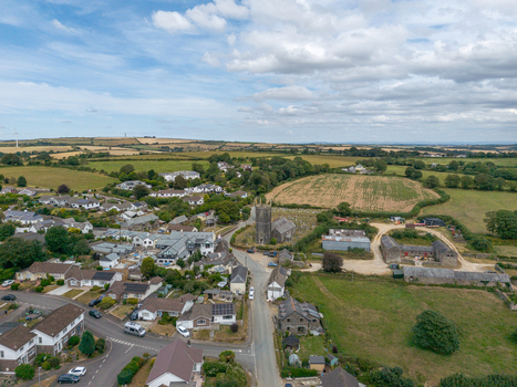 Aerial view of a small village in the countryside featuring a historic stone church residential houses farm buildings and surrounding green fields in Looe Cornwall United Kingdom. Aerial view of a small village in the countryside featuring a historic stone church residential houses farm buildings and surrounding green fields in Looe Cornwall United Kingdom.
