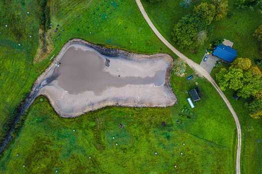 Aerial view of a small pond in a rural landscape surrounded by green fields a dirt road and a house with a blue roof in Latvia.