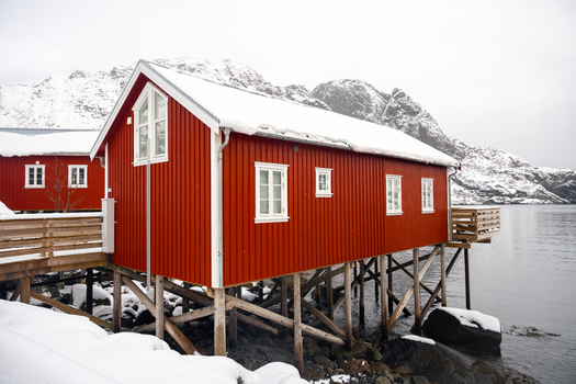 View of traditional red wooden rorbuer cabins on stilts over the water with snow covered mountains in the background in Leknes Lofoten Norway.