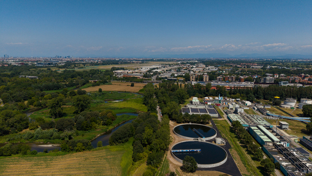 Aerial view of a water treatment plant with circular settling tanks surrounded by lush green fields a winding river and the distant urban skyline of Milan Lombardy Italy. Aerial view of a water treatment plant with circular settling tanks surrounded by lush green fields a winding river and the distant urban skyline of Milan Lombardy Italy.