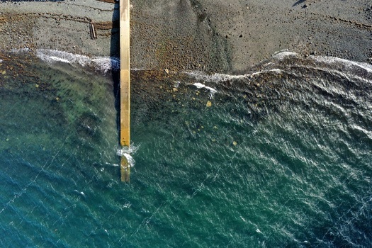 Aerial view of a concrete pier extending into the clear blue waters of the sea with a rocky shoreline and gentle waves in Stein Scotland United Kingdom. Aerial view of a concrete pier extending into the clear blue waters of the sea with a rocky shoreline and gentle waves in Stein Scotland United Kingdom.