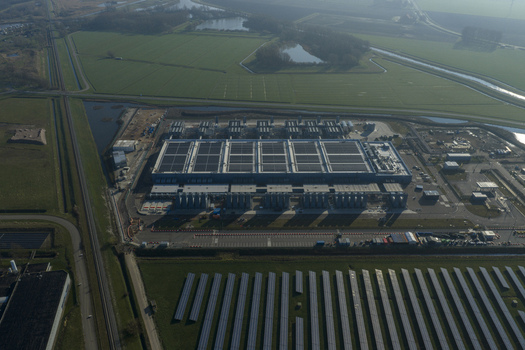 Aerial view of an AI data center with cooling towers and a cloud computing hub exterior featuring a blue industrial building solar panels and green fields. Aerial view of an AI data center with cooling towers and a cloud computing hub exterior featuring a blue industrial building solar panels and green fields.