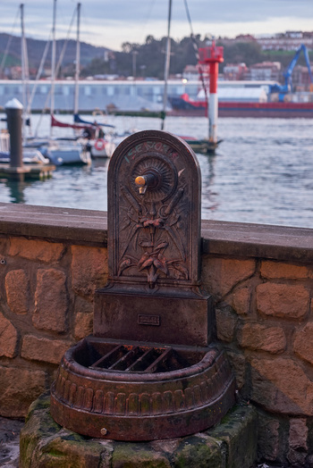 Pasai Donibane Spain 09 March 2026 View of an ornate cast iron water fountain on a stone wall overlooking the harbor with boats and a red beacon in the background.