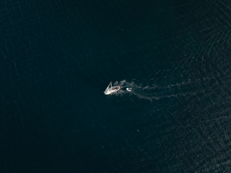 Aerial view of a white sailboat with a small dinghy in tow cruising across the deep blue sea near the Island of Ortigia Syracuse Sicily Italy. Aerial view of a white sailboat with a small dinghy in tow cruising across the deep blue sea near the Island of Ortigia Syracuse Sicily Italy.