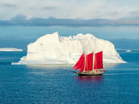 Aerial view of a sailboat with vibrant red sails navigating through deep blue Arctic waters past a massive white iceberg under a clear sky in Greenland. Aerial view of a sailboat with vibrant red sails navigating through deep blue Arctic waters past a massive white iceberg under a clear sky in Greenland.