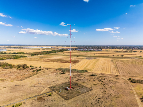 Aerial view of a tall red and white telecommunications tower standing in a vast field of dry grass under a clear blue sky in Szigetszentmiklos Pest County Hungary. Aerial view of a tall red and white telecommunications tower standing in a vast field of dry grass under a clear blue sky in Szigetszentmiklos Pest County Hungary.