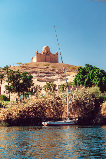 View of the Mausoleum of the Aga Khan on a desert hill with a traditional felucca boat on the Nile river in Aswan Aswan Governorate Egypt. View of the Mausoleum of the Aga Khan on a desert hill with a traditional felucca boat on the Nile river in Aswan Aswan Governorate Egypt.