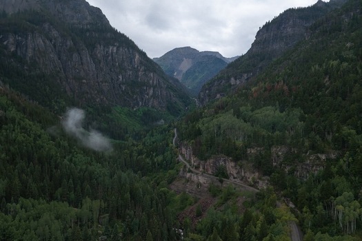 Aerial view of the rugged Yankee Boy Basin near Mt. Sneffels featuring dense forests a winding mountain road and rocky peaks under a cloudy sky in Ouray Colorado United States.