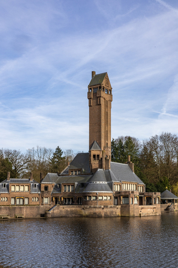 View of Country Residence Museum Jachthuis Sint Hubertus with its iconic tall brick tower reflecting in the calm water under a blue sky Hoenderloo Gelderland Netherlands.