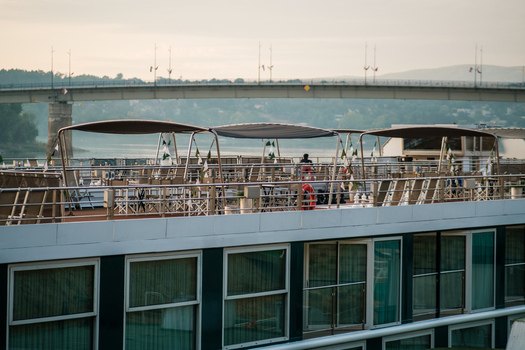 View of the deck of a river cruise ship with sunshades outdoor seating and a lifebuoy with a bridge spanning the river in the background in Novi Sad Vojvodina Serbia. View of the deck of a river cruise ship with sunshades outdoor seating and a lifebuoy with a bridge spanning the river in the background in Novi Sad Vojvodina Serbia.