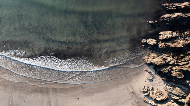 Aerial view of gentle waves rolling onto a sandy beach and jagged coastal rocks in Mothecombe Devon United Kingdom. Aerial view of gentle waves rolling onto a sandy beach and jagged coastal rocks in Mothecombe Devon United Kingdom.