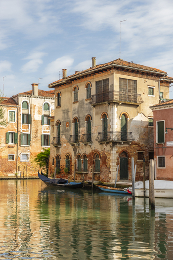 View of historic Venetian buildings with weathered facades and balconies along a canal with a gondola and small boats in Venice Veneto Italy. View of historic Venetian buildings with weathered facades and balconies along a canal with a gondola and small boats in Venice Veneto Italy.