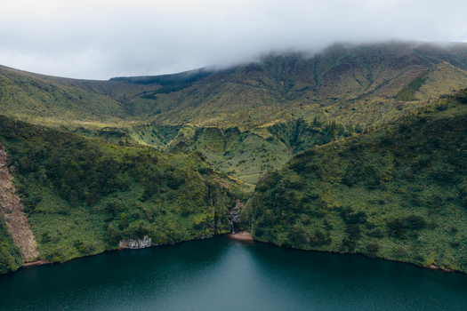 Aerial view of Caldeira Funda crater lake with a small waterfall cascading into dark blue water surrounded by lush green volcanic slopes under a misty sky Lajes das Flores Azores Portugal. Aerial view of Caldeira Funda crater lake with a small waterfall cascading into dark blue water surrounded by lush green volcanic slopes under a misty sky Lajes das Flores Azores Portugal.