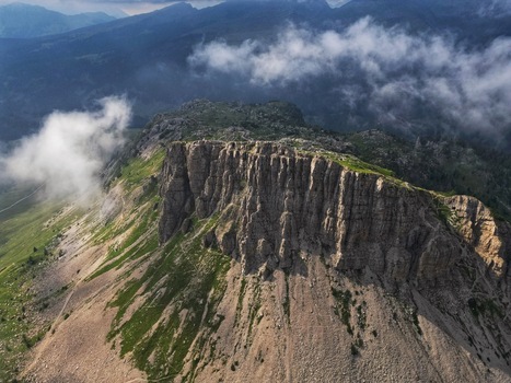 Aerial view of the Dolomite Mountains featuring steep rocky cliffs and green slopes under a misty sky in Canazei Trentino South Tyrol Italy. Aerial view of the Dolomite Mountains featuring steep rocky cliffs and green slopes under a misty sky in Canazei Trentino South Tyrol Italy.