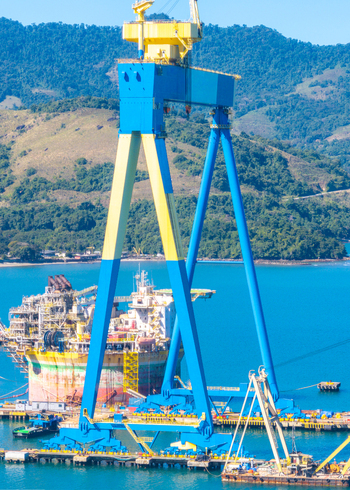 Aerial view of a large blue and yellow gantry crane at a shipyard with an offshore oil platform vessel surrounded by coastal hills and blue water in Angra dos Reis Rio de Janeiro Brazil.