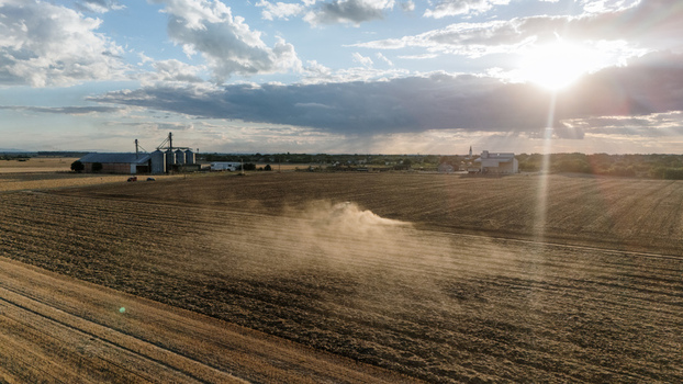 Aerial view of a tractor working in a vast agricultural field kicking up dust during golden hour with grain silos in the background in Veliki Radinci Vojvodina Serbia. Aerial view of a tractor working in a vast agricultural field kicking up dust during golden hour with grain silos in the background in Veliki Radinci Vojvodina Serbia.