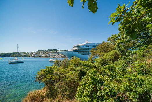 View of a large cruise ship anchored in the Fowey River near the town of Fowey with sailboats and lush trees in the foreground under a clear blue sky in Fowey Cornwall United Kingdom. View of a large cruise ship anchored in the Fowey River near the town of Fowey with sailboats and lush trees in the foreground under a clear blue sky in Fowey Cornwall United Kingdom.