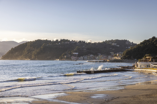 View of Venere Azzurra beach with waves crashing against a pier and lush green hills under a clear sky in Lerici Liguria Italy.