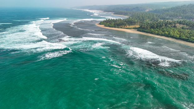 Aerial view of the turquoise ocean waves crashing onto a sandy beach lined with lush palm trees and tropical vegetation along the coastline of Indonesia. Aerial view of the turquoise ocean waves crashing onto a sandy beach lined with lush palm trees and tropical vegetation along the coastline of Indonesia.
