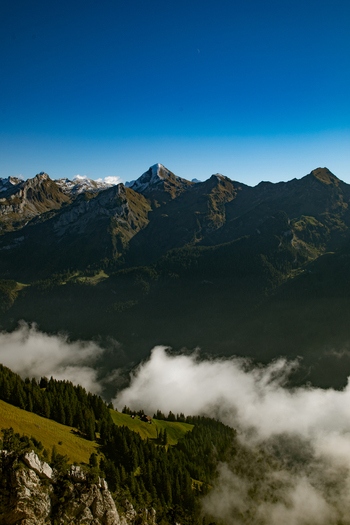 Aerial view of the Swiss Alps with snow capped mountain peaks lush green slopes dense pine forests and low hanging clouds under a clear blue sky in Airolo Ticino Switzerland. Aerial view of the Swiss Alps with snow capped mountain peaks lush green slopes dense pine forests and low hanging clouds under a clear blue sky in Airolo Ticino Switzerland.