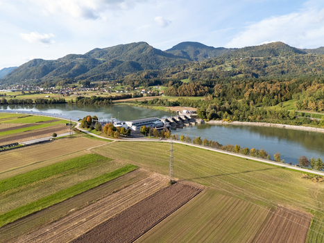 Aerial view of a hydroelectric power plant on a river with agricultural fields and power lines in the foreground and forested mountains in the background in Slovenia.