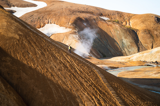 View of a hiker in a yellow jacket walking up a wooden staircase on the steep orange brown volcanic slopes of Kerlingarfjoll with geothermal steam in Hrunamannahreppur Iceland.