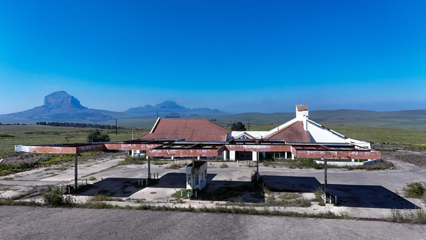 Aerial view of an abandoned gas station with a rusted canopy and red roofed building set against a backdrop of flat plains and distant mountains under a clear blue sky in iNtabazwe Free State South Africa. Aerial view of an abandoned gas station with a rusted canopy and red roofed building set against a backdrop of flat plains and distant mountains under a clear blue sky in iNtabazwe Free State South Africa.