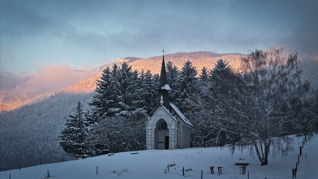 Aerial view of a small stone chapel surrounded by snow covered pine trees and mountains during golden hour in Vesancy Auvergne Rhone Alpes France. Aerial view of a small stone chapel surrounded by snow covered pine trees and mountains during golden hour in Vesancy Auvergne Rhone Alpes France.