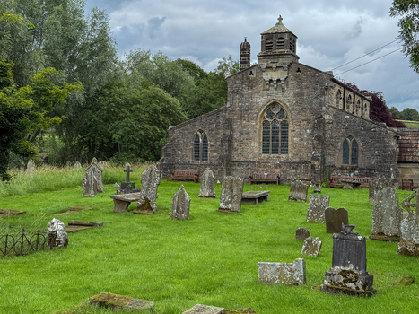 View of the historic stone church and graveyard with weathered headstones and lush green trees under an overcast sky in Hawes England United Kingdom. View of the historic stone church and graveyard with weathered headstones and lush green trees under an overcast sky in Hawes England United Kingdom.