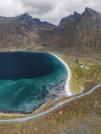 Aerial view of Senja island featuring a turquoise bay dramatic rugged mountains a winding coastal road and small houses along the shoreline in Senja Troms Norway.