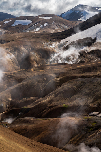 View of geothermal steam rises from the ochre and umber earth contrasting with the distant snow capped mountains under a dramatic sky Hrunamannahreppur Iceland.