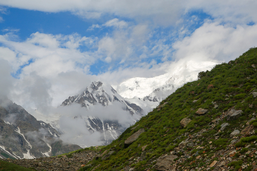 View of the snow capped peaks of Rama Meadows rising above lush green slopes and rocky terrain under a cloudy blue sky in Astore Gilgit Pakistan. View of the snow capped peaks of Rama Meadows rising above lush green slopes and rocky terrain under a cloudy blue sky in Astore Gilgit Pakistan.