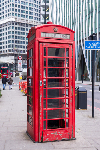 London United Kingdom 12 May 2025 View of a classic red telephone box on a city sidewalk with modern architecture and pedestrians in the background.