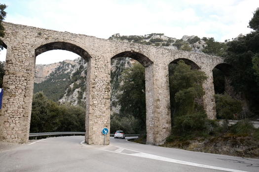 View of an ancient stone aqueduct with multiple arches spanning over a winding mountain road in Mallorca Balearic Islands Spain.