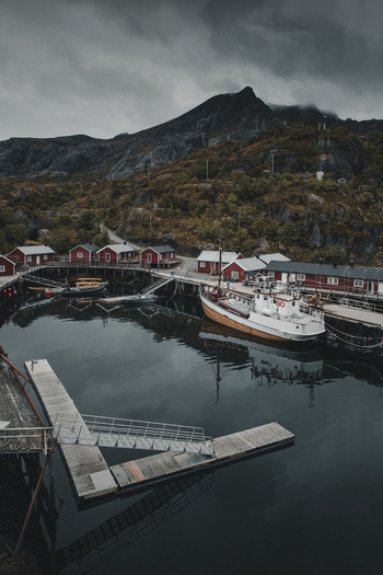 Nusfjord Norway 07 September 2022 View of the historic fishing village of Nusfjord with iconic red wooden cabins a white fishing boat at the pier and dramatic mountains. Nusfjord Norway 07 September 2022 View of the historic fishing village of Nusfjord with iconic red wooden cabins a white fishing boat at the pier and dramatic mountains.