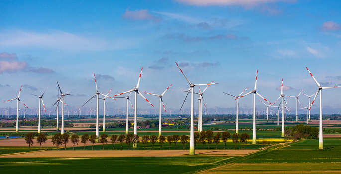 Aerial view of a large wind farm with numerous white wind turbines set against a vast green agricultural landscape under a clear blue sky in Germany. Aerial view of a large wind farm with numerous white wind turbines set against a vast green agricultural landscape under a clear blue sky in Germany.