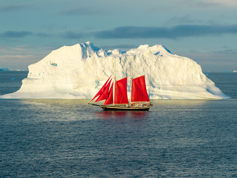 Aerial view of a sailboat with striking red sails navigating the cold blue waters in front of a massive white iceberg under a clear sky in Greenland. Aerial view of a sailboat with striking red sails navigating the cold blue waters in front of a massive white iceberg under a clear sky in Greenland.