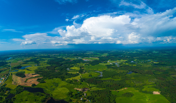 Aerial view of lush green fields dense forests and small lakes under a vast blue sky with large white clouds in Latvia. Aerial view of lush green fields dense forests and small lakes under a vast blue sky with large white clouds in Latvia.