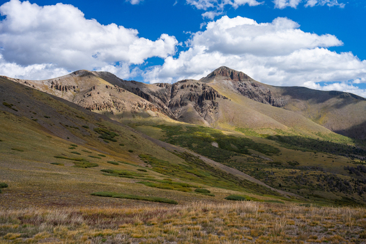 View of the rugged slopes and rocky summit of San Luis Peak under a bright blue sky with scattered clouds featuring alpine tundra and mountain terrain near Creede Colorado United States. View of the rugged slopes and rocky summit of San Luis Peak under a bright blue sky with scattered clouds featuring alpine tundra and mountain terrain near Creede Colorado United States.