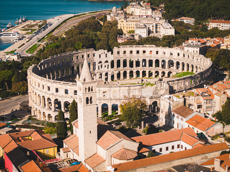 Aerial view of the majestic Pula Arena stands in ancient splendor against the backdrop of the Adriatic Sea framed by terracotta rooftops and lush greenery Pula Istria County Croatia.
