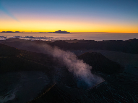 Aerial view of Mount Bromo emitting volcanic smoke at sunrise with a sea of clouds and distant mountain peaks in Tosari East Java Indonesia. Aerial view of Mount Bromo emitting volcanic smoke at sunrise with a sea of clouds and distant mountain peaks in Tosari East Java Indonesia.
