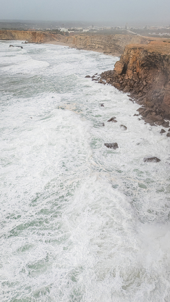 Aerial view of the rugged cliffs with powerful waves crashing against the rocky shoreline under a misty sky in Sagres Faro Portugal. Aerial view of the rugged cliffs with powerful waves crashing against the rocky shoreline under a misty sky in Sagres Faro Portugal.