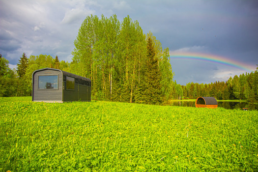 View of a modern black cabin situated in a lush green field with dandelions a pond and a rainbow arching over the forest in Latvia. View of a modern black cabin situated in a lush green field with dandelions a pond and a rainbow arching over the forest in Latvia.